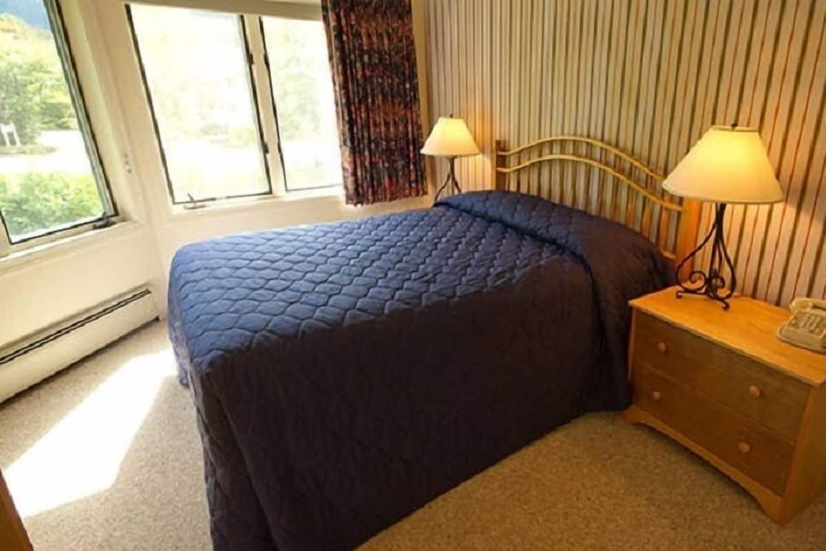 Um quarto com uma cama, duas luminárias de cabeceira, uma mesinha de cabeceira com telefone e uma janela com cortinas.