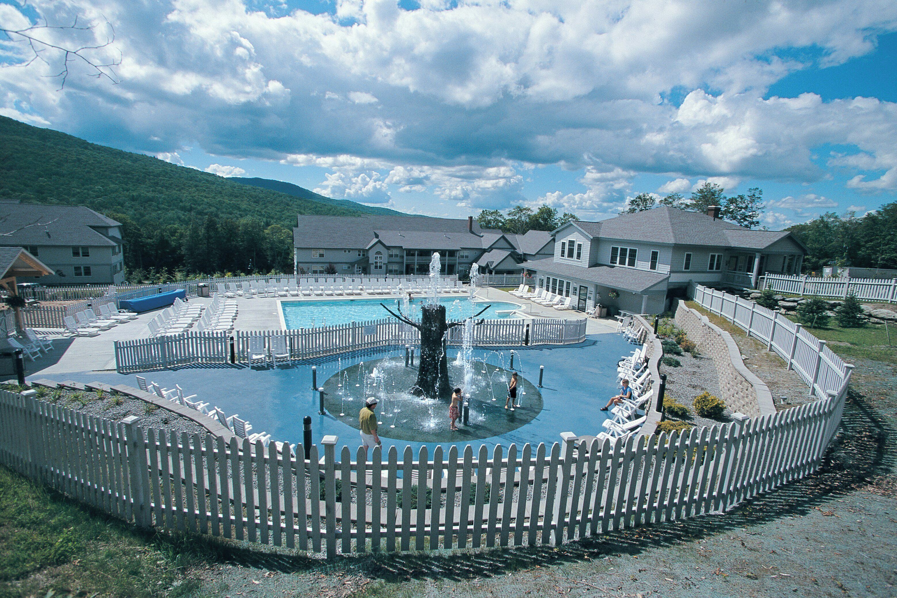 A swimming pool with a fountain, surrounded by a white picket fence, with houses in the background.