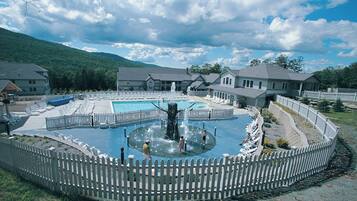 A swimming pool with a fountain, surrounded by a white picket fence, with houses in the background.