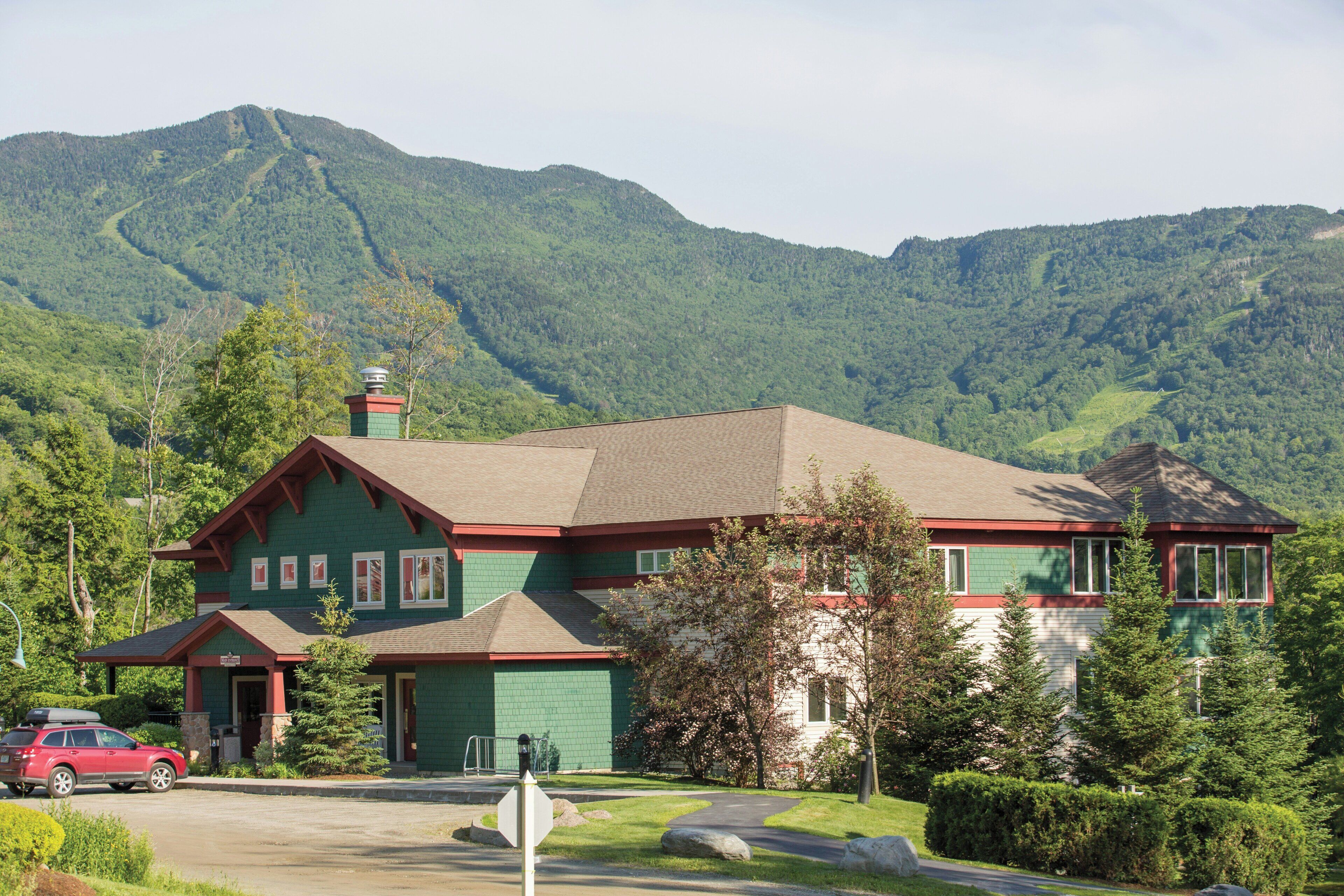 Edificio de dos plantas con fachada verde y detalles en rojo, rodeado de abundante vegetación y montañas al fondo.