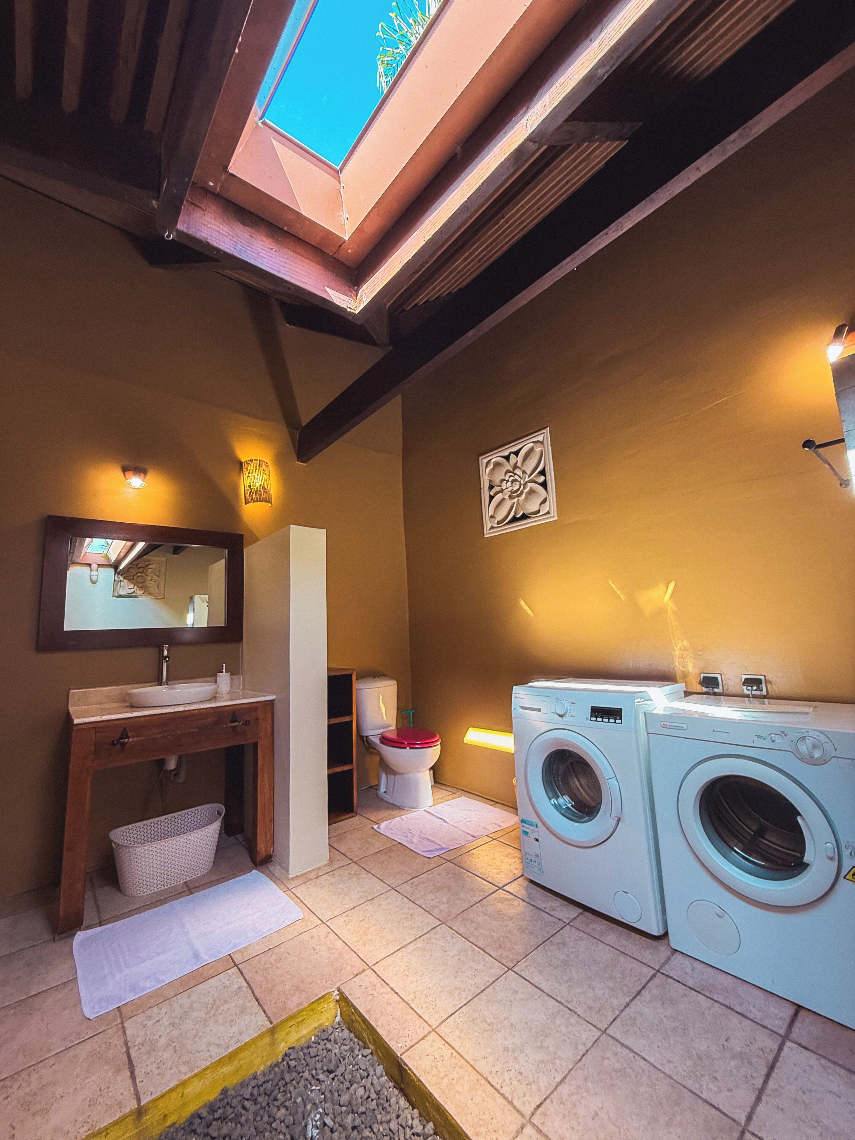 A compact bathroom with a sink, toilet, and washing machine, featuring a skylight and tiled flooring.