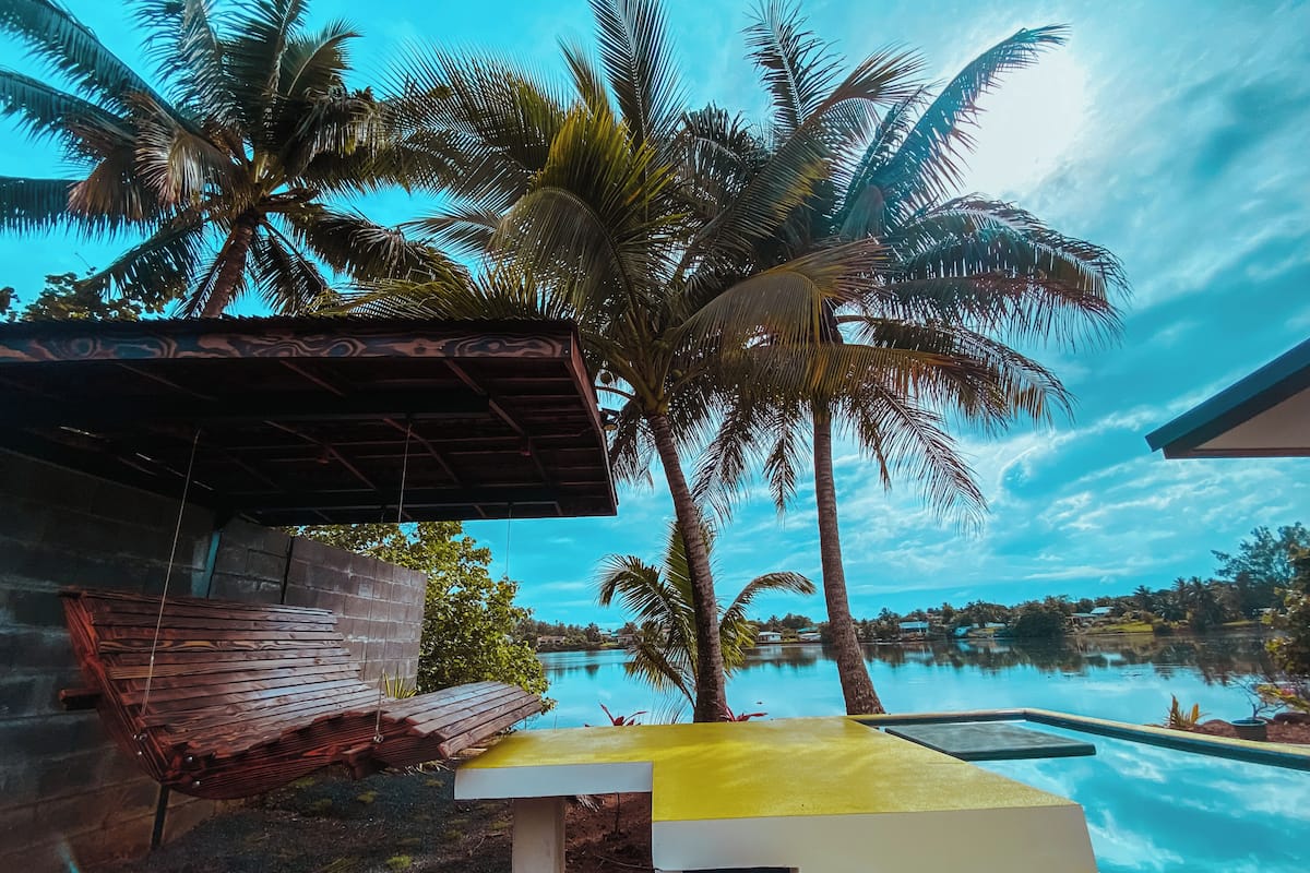 Zona junto a la piscina con un banco, palmeras y vista a un cuerpo de agua.