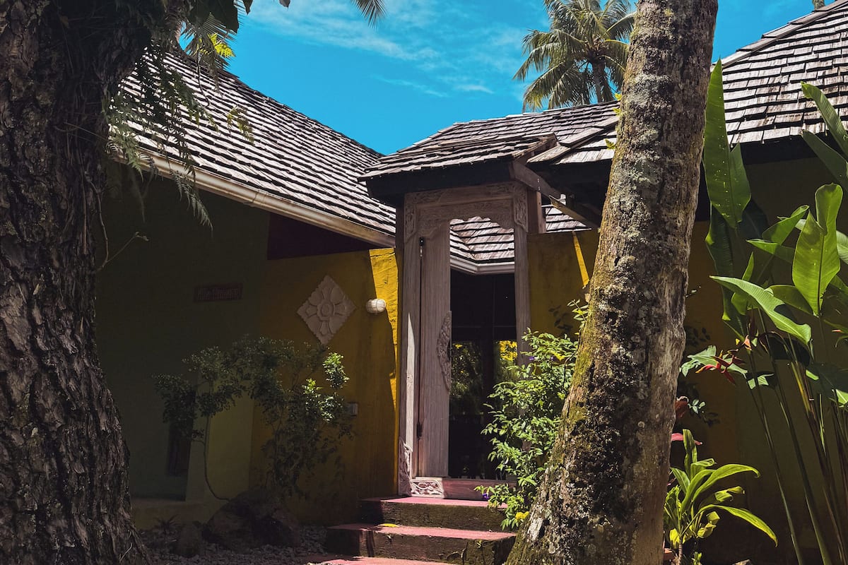 Un entorno tropical con un edificio que tiene techo de tejas, un porche y escalones que llevan a una puerta.