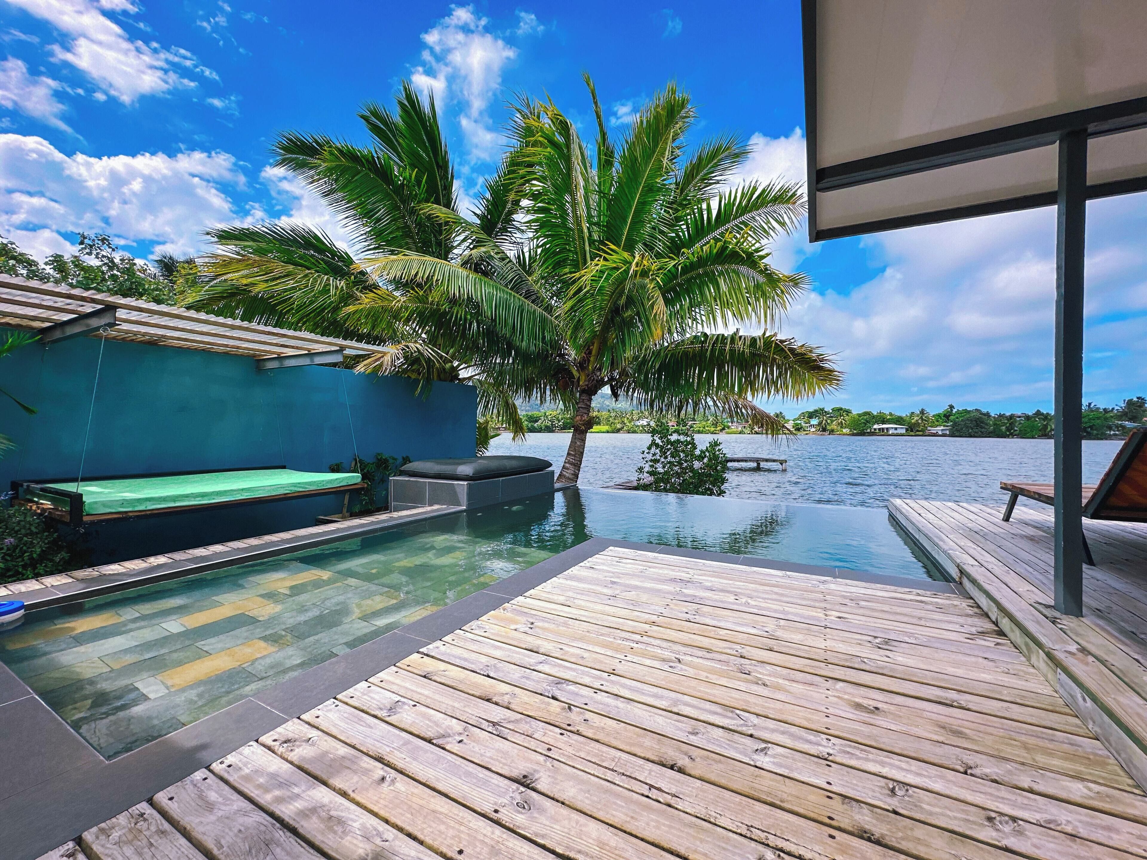 Una piscina infinita con cubierta de madera, una palmera y vista al agua.