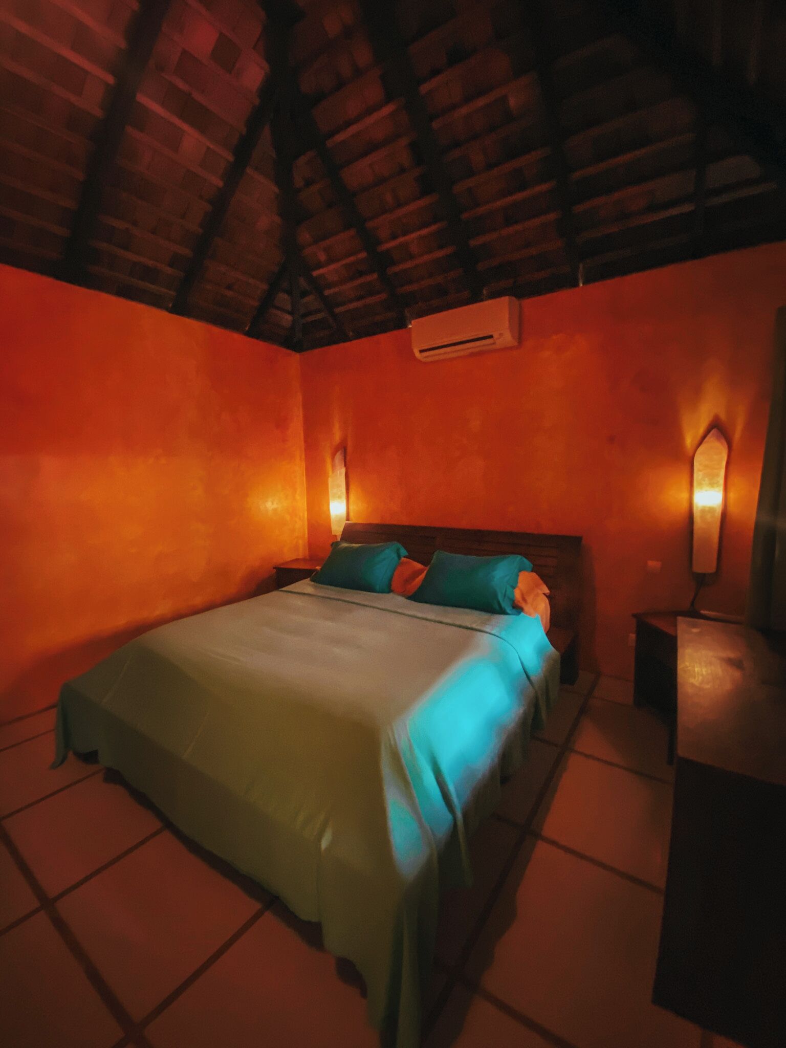 A bedroom with a bed, a nightstand, an air conditioning unit, and a wall-mounted lamp.