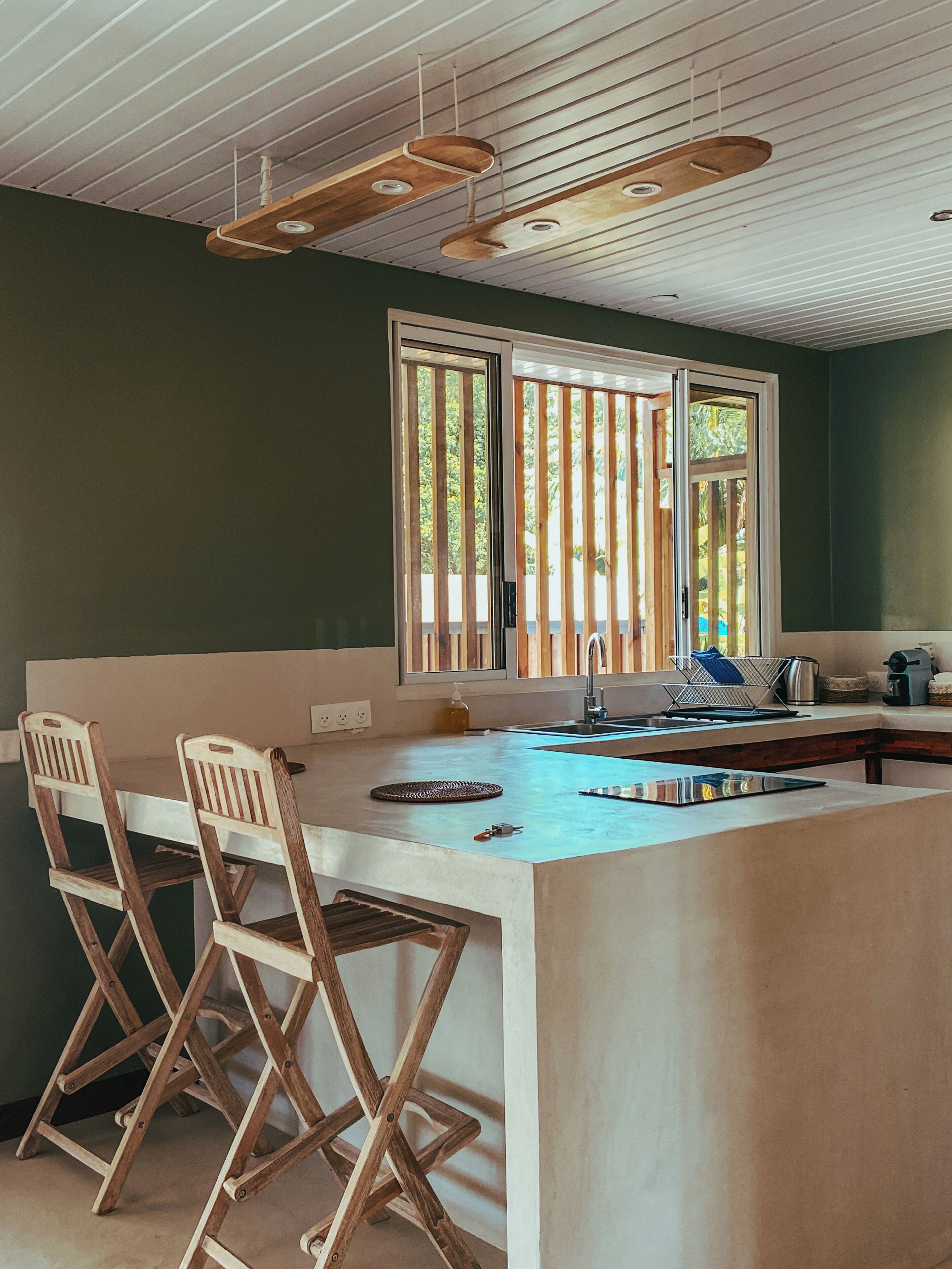 A kitchen with a large island, wooden chairs, a window with vertical blinds, and pendant lights.