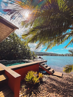 A pool area with a wooden deck, lounge chairs, and a view of a lake. - Villa Mitirapa (Taiarapu-Ouest)