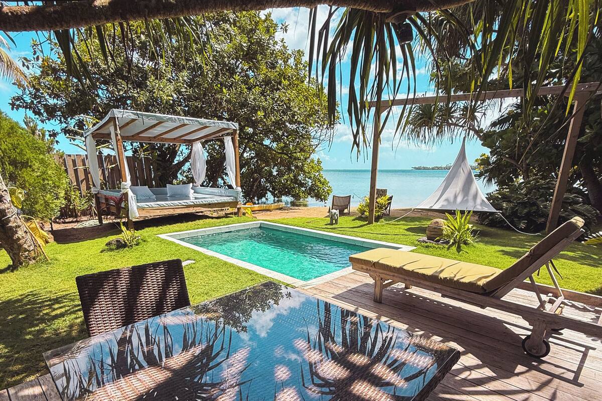 Zona junto a la piscina con mesa de vidrio, sillas de mimbre, una pérgola y vistas al mar.