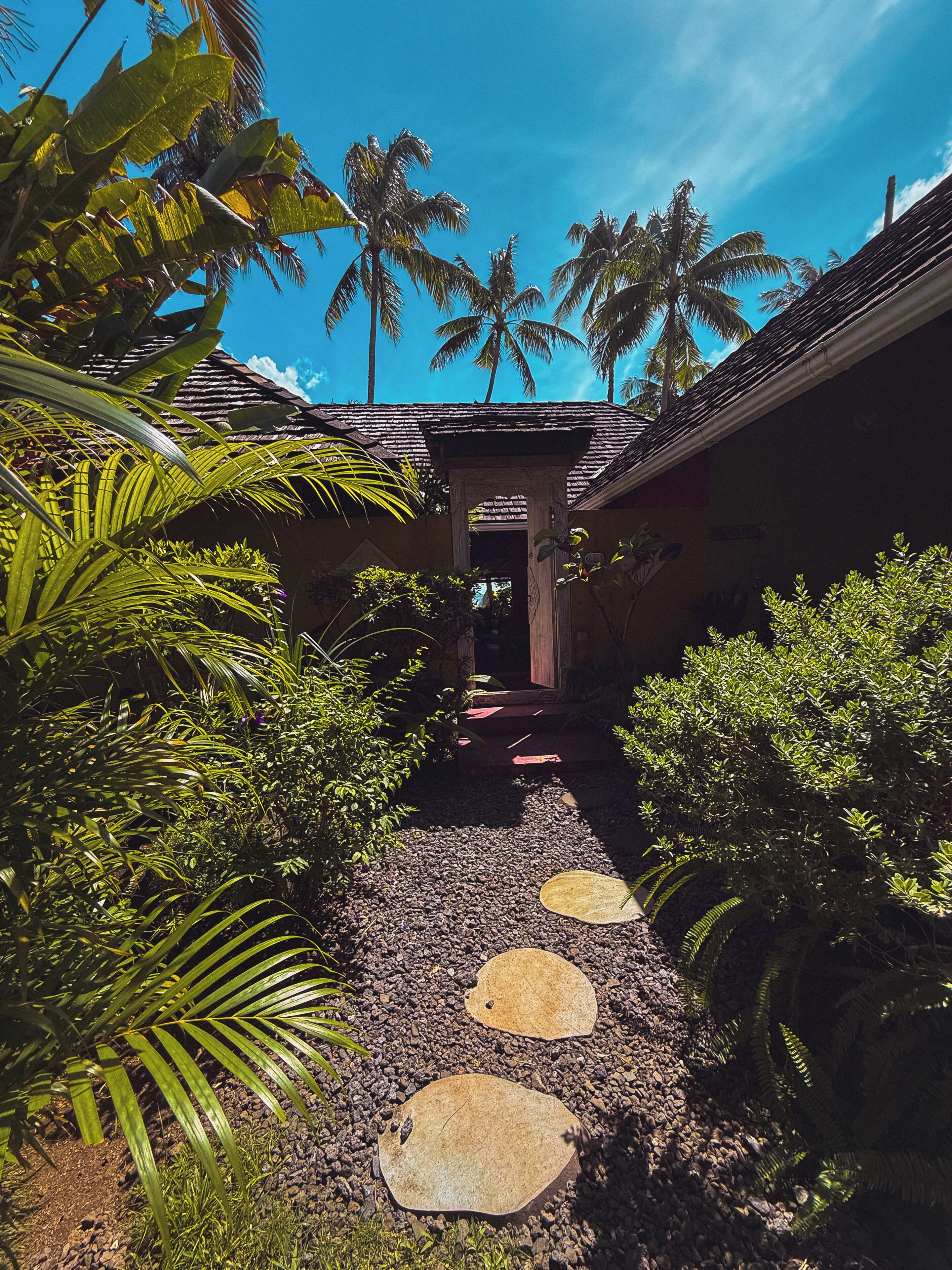 Un chemin menant à un bâtiment doté d’un toit en tuiles, entouré d’une végétation tropicale et de palmiers.