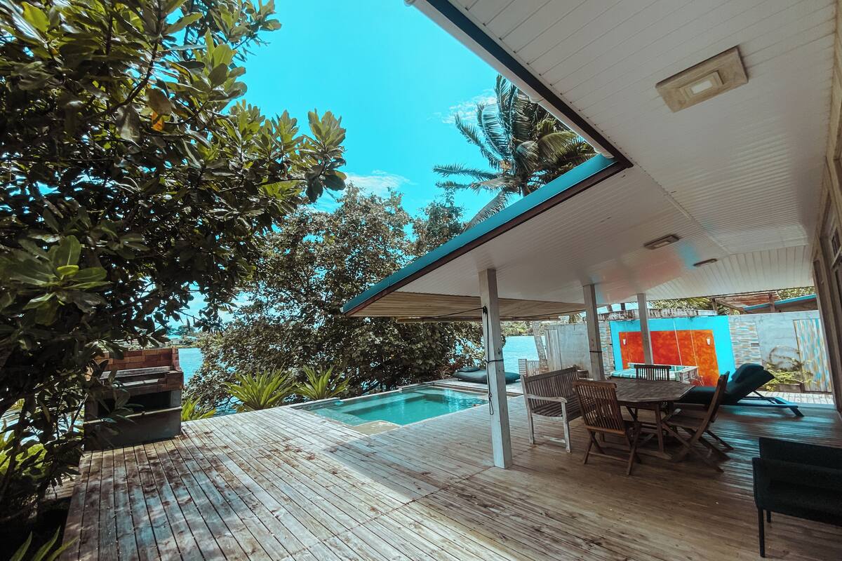 Una terraza de madera con piscina, mobiliario exterior y una zona cubierta con vista al mar.