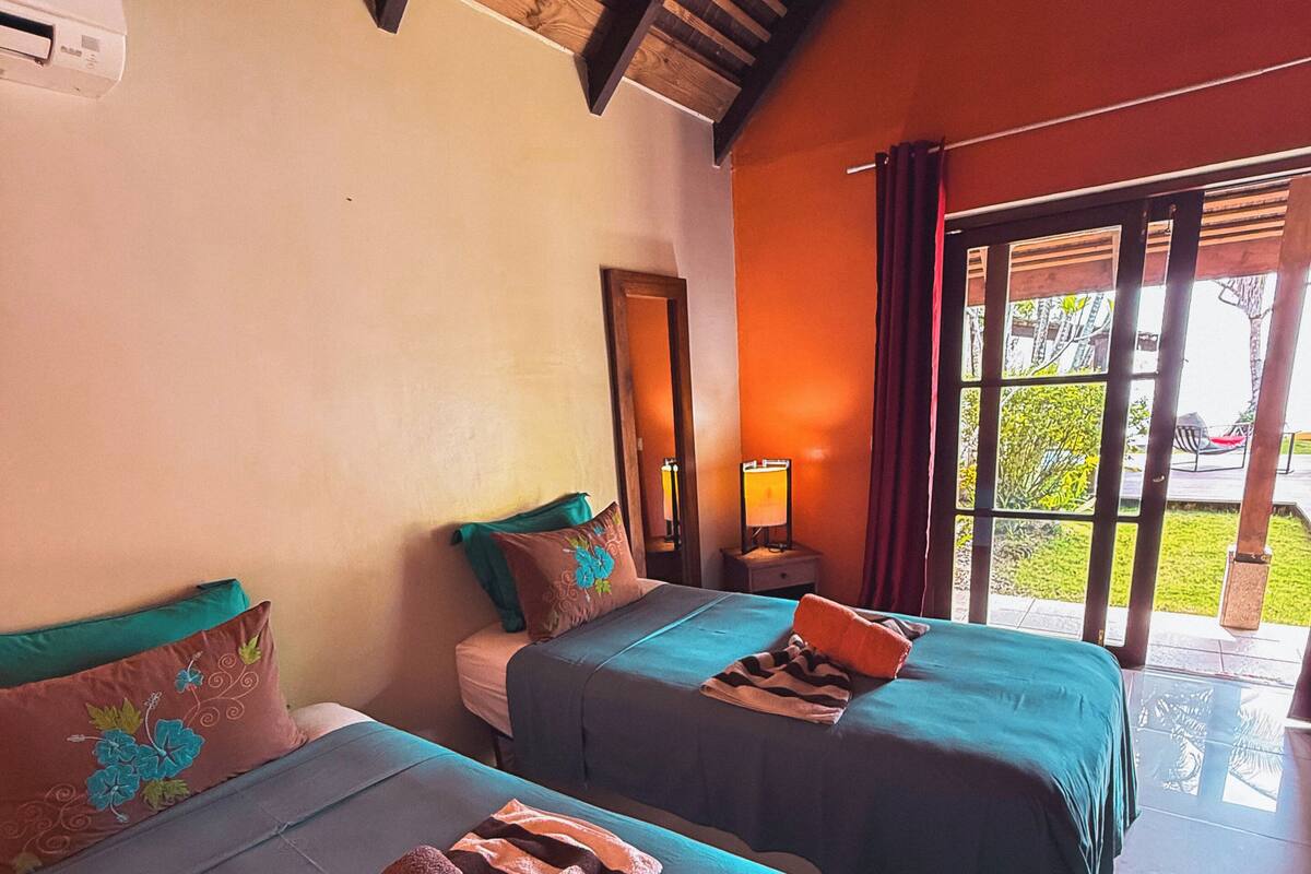 Una habitación de hotel con dos camas, un espejo, una mesita de noche con lámpara y un ventanal corredero que da a un espacio exterior.