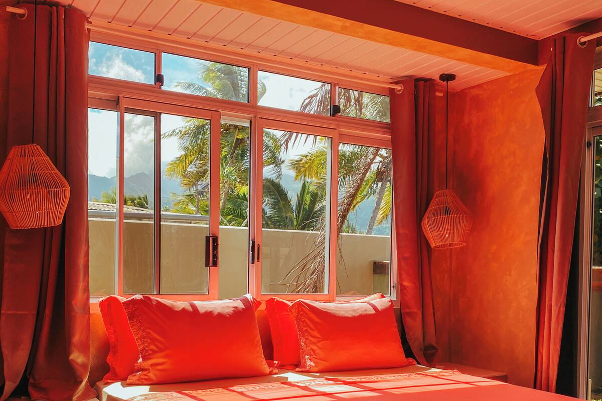 Un dormitorio con una cama grande, cortinas rojas y vistas a palmeras y montañas a través del ventanal.