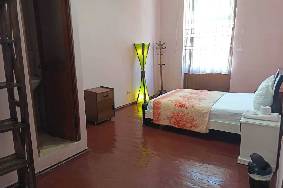Quarto com cama, mesa de cabeceira, armário de madeira pequeno, luminária de chão e janela com cortinas.