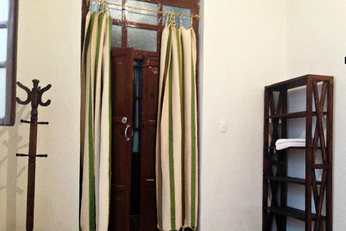 Um quarto com um porta-roupas de madeira, uma porta com cortinas verdes e brancas, um tapete estampado e uma estante de madeira.