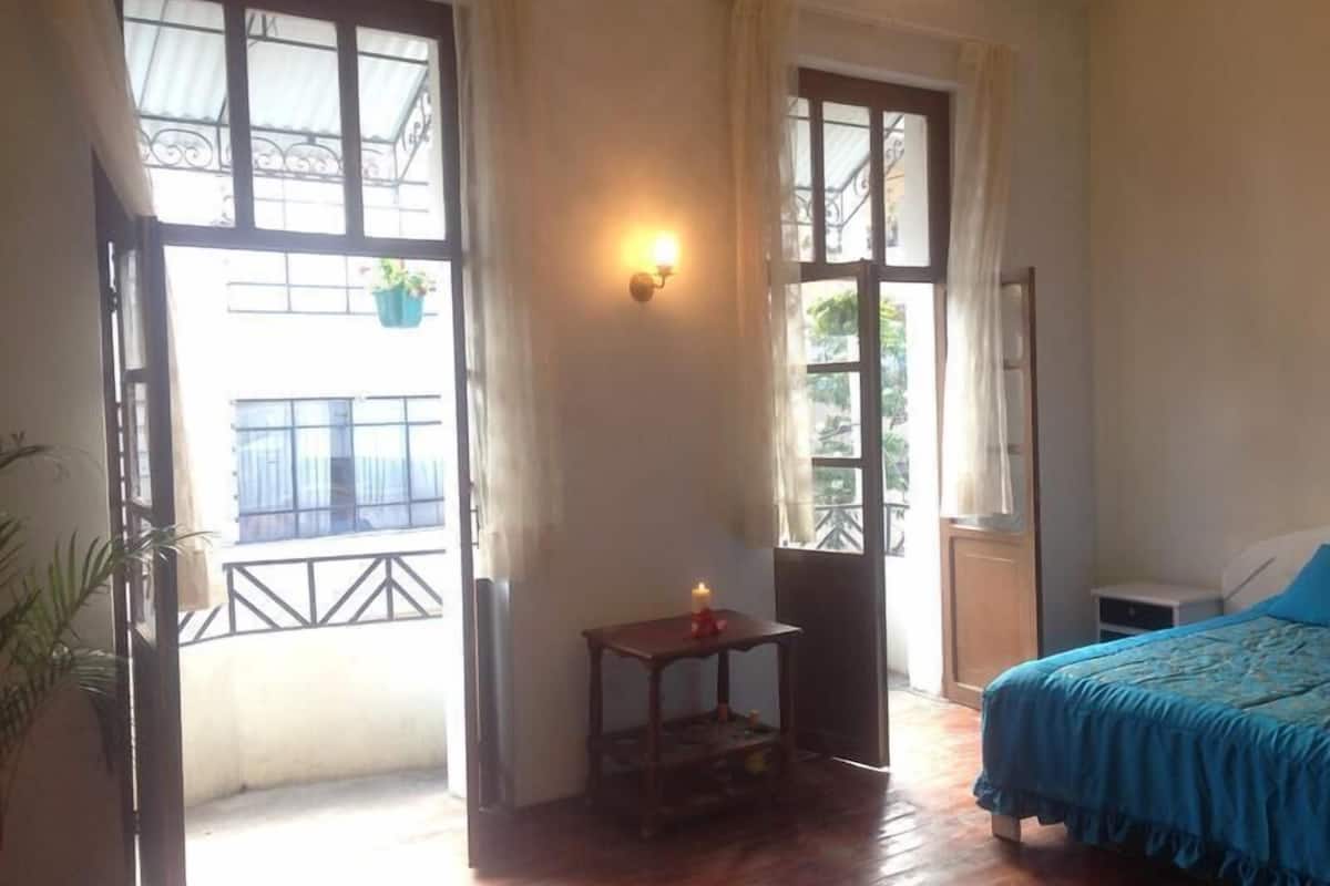 Quarto com cama, uma pequena mesa e janela com vista para uma varanda.