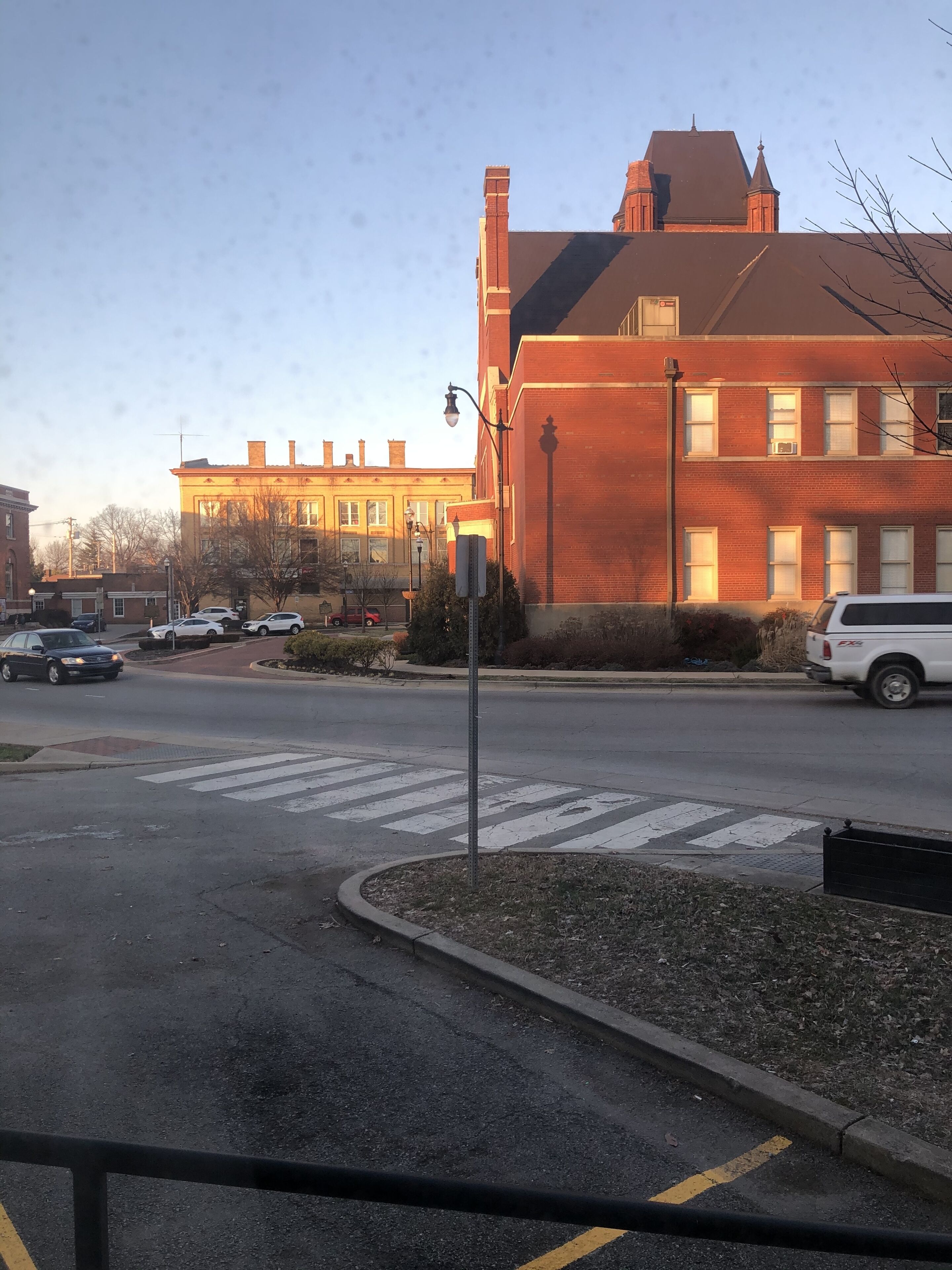 Vista de una calle con un paso de peatones, un SUV blanco y un edificio de ladrillo con varias ventanas y una chimenea.