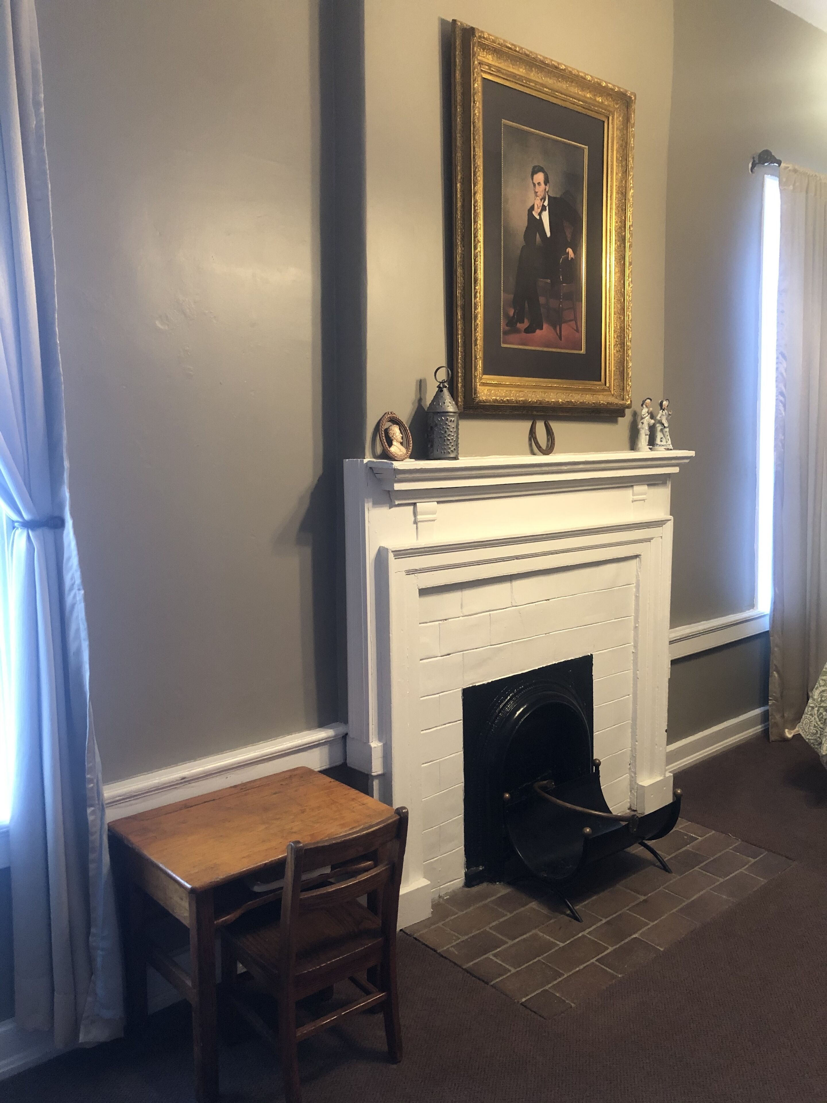 Una habitación con chimenea blanca, un retrato enmarcado encima, una mesita con una silla y una ventana con cortinas.