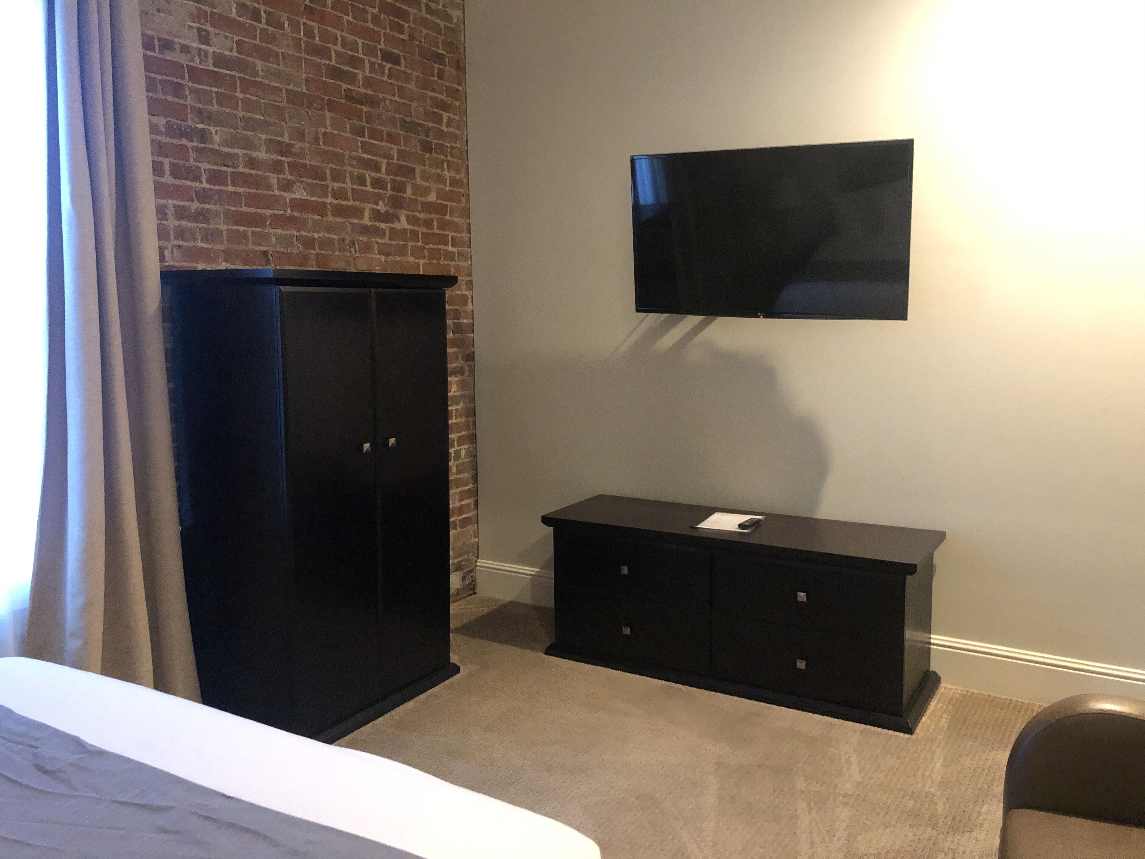 A room with a brick wall, a black wardrobe, a flat-screen TV mounted on the wall, and a small table with a remote control.