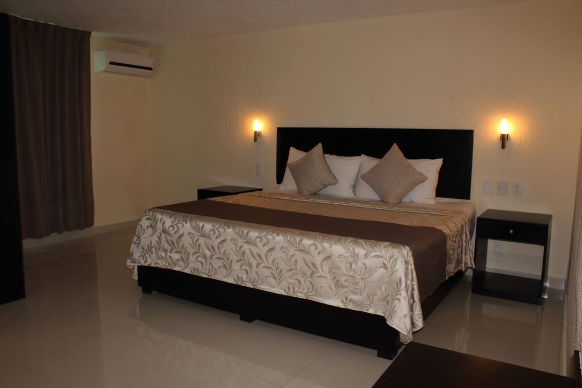 A hotel room with a large bed, two bedside tables, an air conditioning unit, and wall-mounted lights.