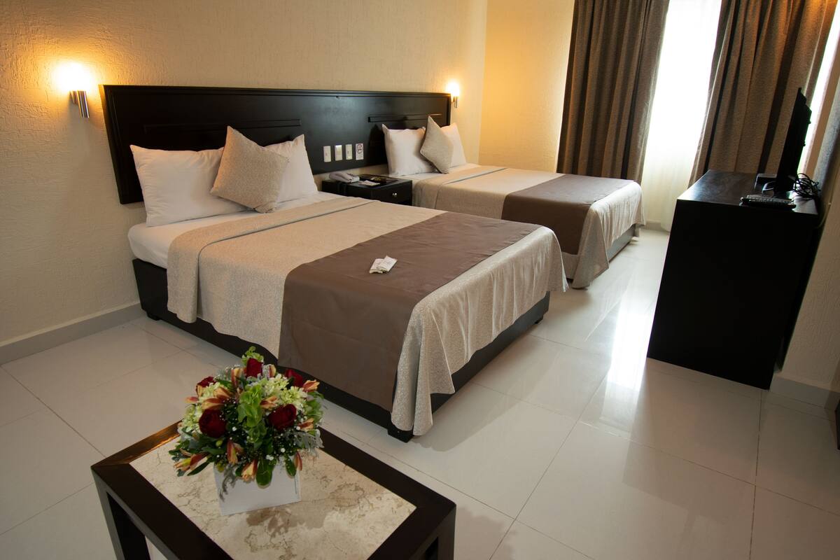 A hotel room with two beds, a nightstand, a TV stand, and a floral centerpiece on a table.