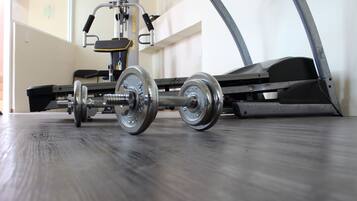 A weight machine with adjustable weights and a bench, positioned on a wooden floor in a room with a plain wall and a door in the background.
