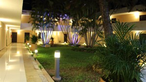 Un pasillo de hotel bien iluminado con un diseño moderno, con entrada por puerta de vidrio, un camino bordeado de pequeñas luces y un área de jardín ajardinado con palmeras y arbustos.
