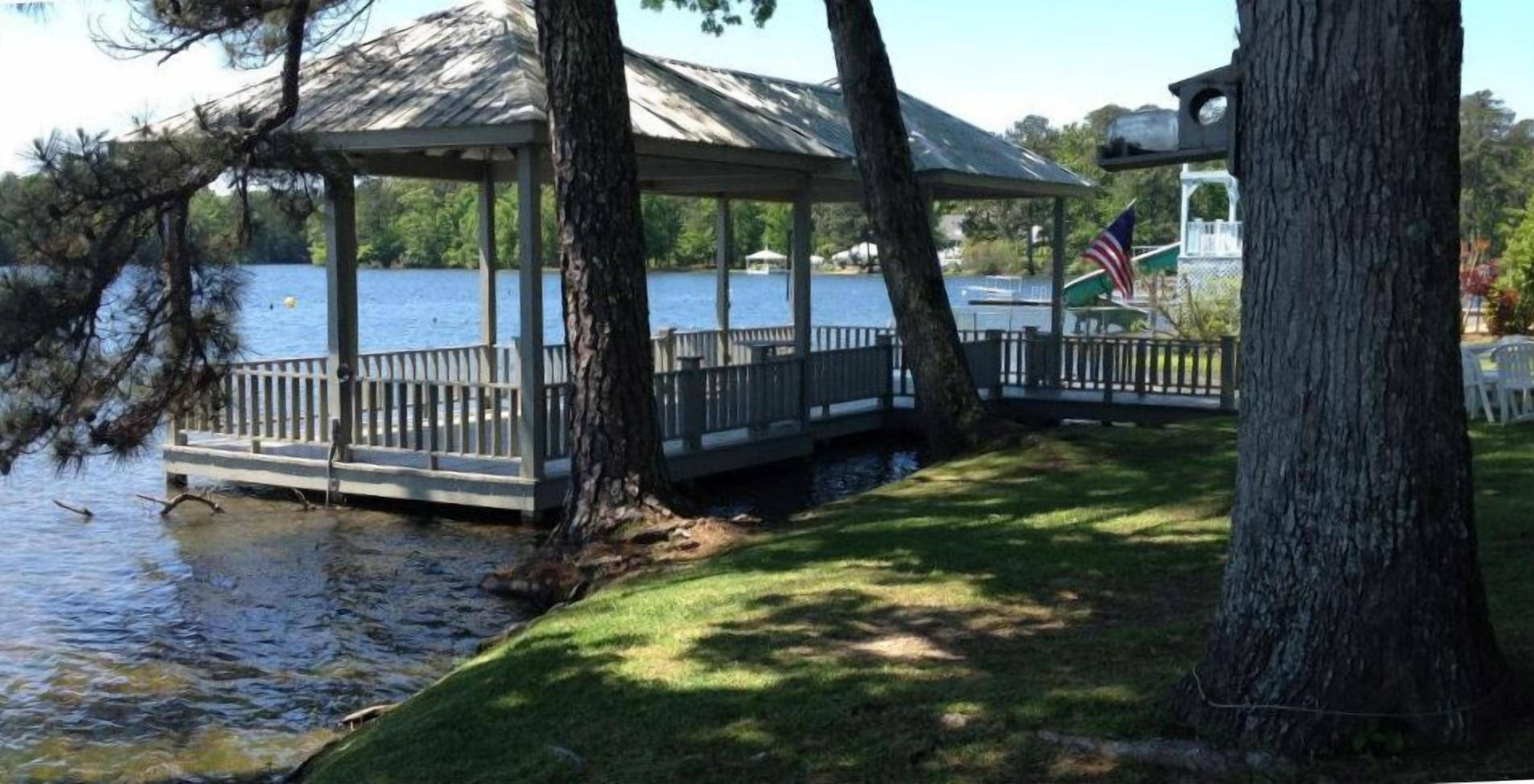 Um gazebo com um deck de madeira com vista para um lago, rodeado por árvores e uma bandeira.