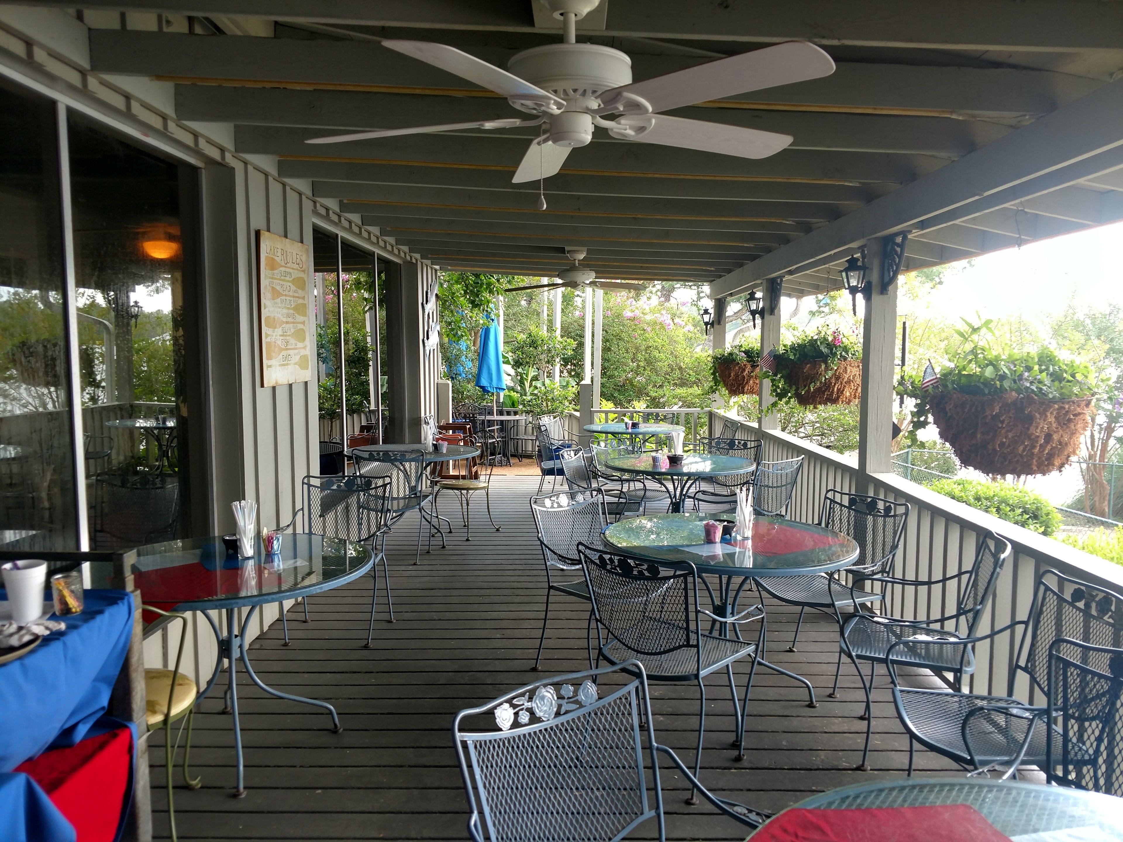 Une terrasse extérieure couverte, dotée de tables et de chaises, d’un ventilateur de plafond et de jardinières suspendues.