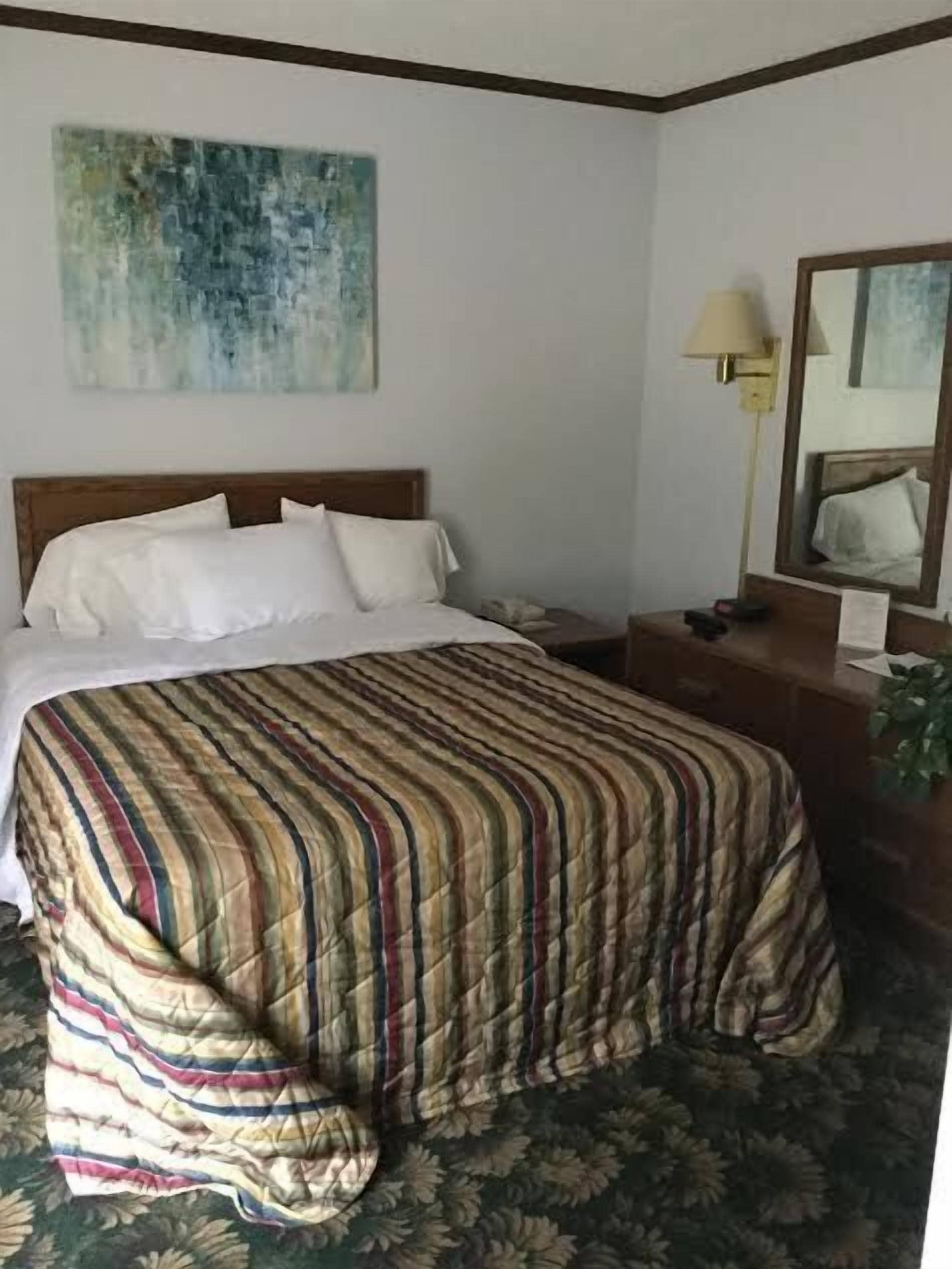 Hotellihuone, jossa on sänky, yöpöytä, lamppu, peili ja kukkakuviollinen matto.