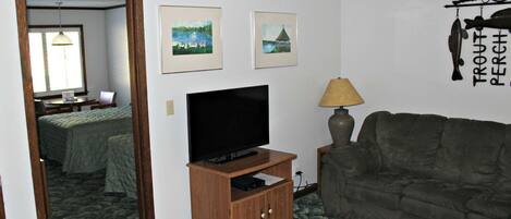 Ein Zimmer mit Fernseher auf einem Holzboden, einer Couch, zwei gerahmten Bildern an der Wand und einem kleinen Tisch mit einer Lampe.