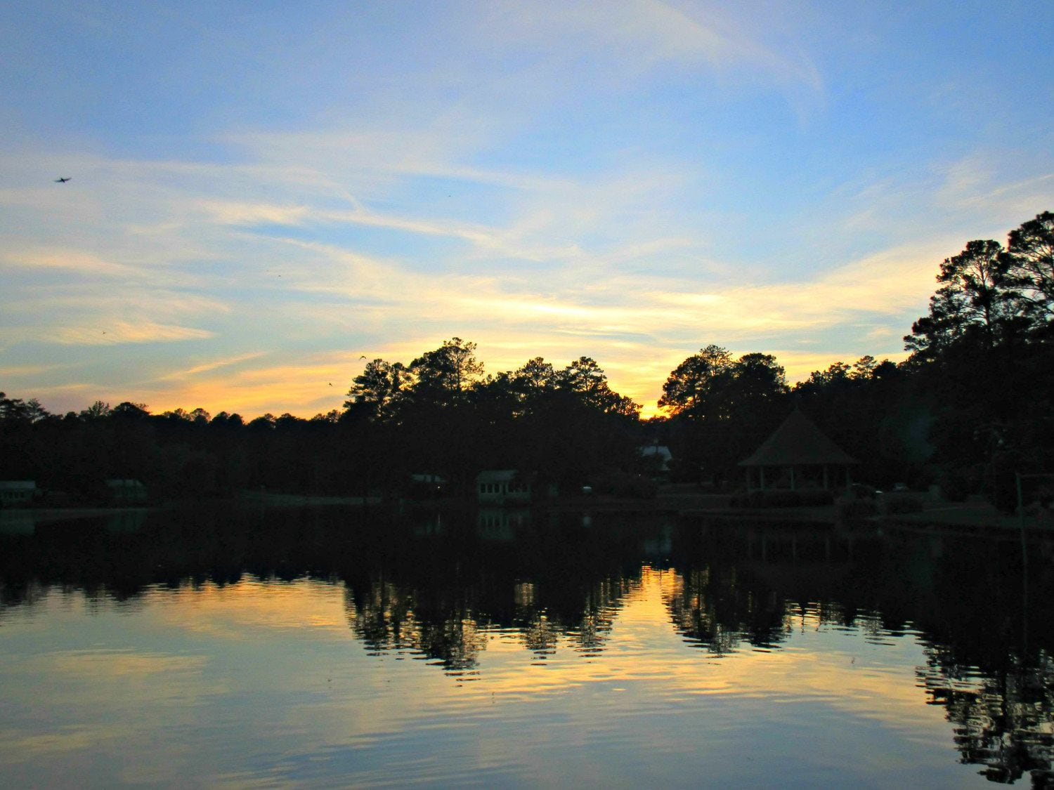 A calm lake at sunset with reflections of trees and buildings.