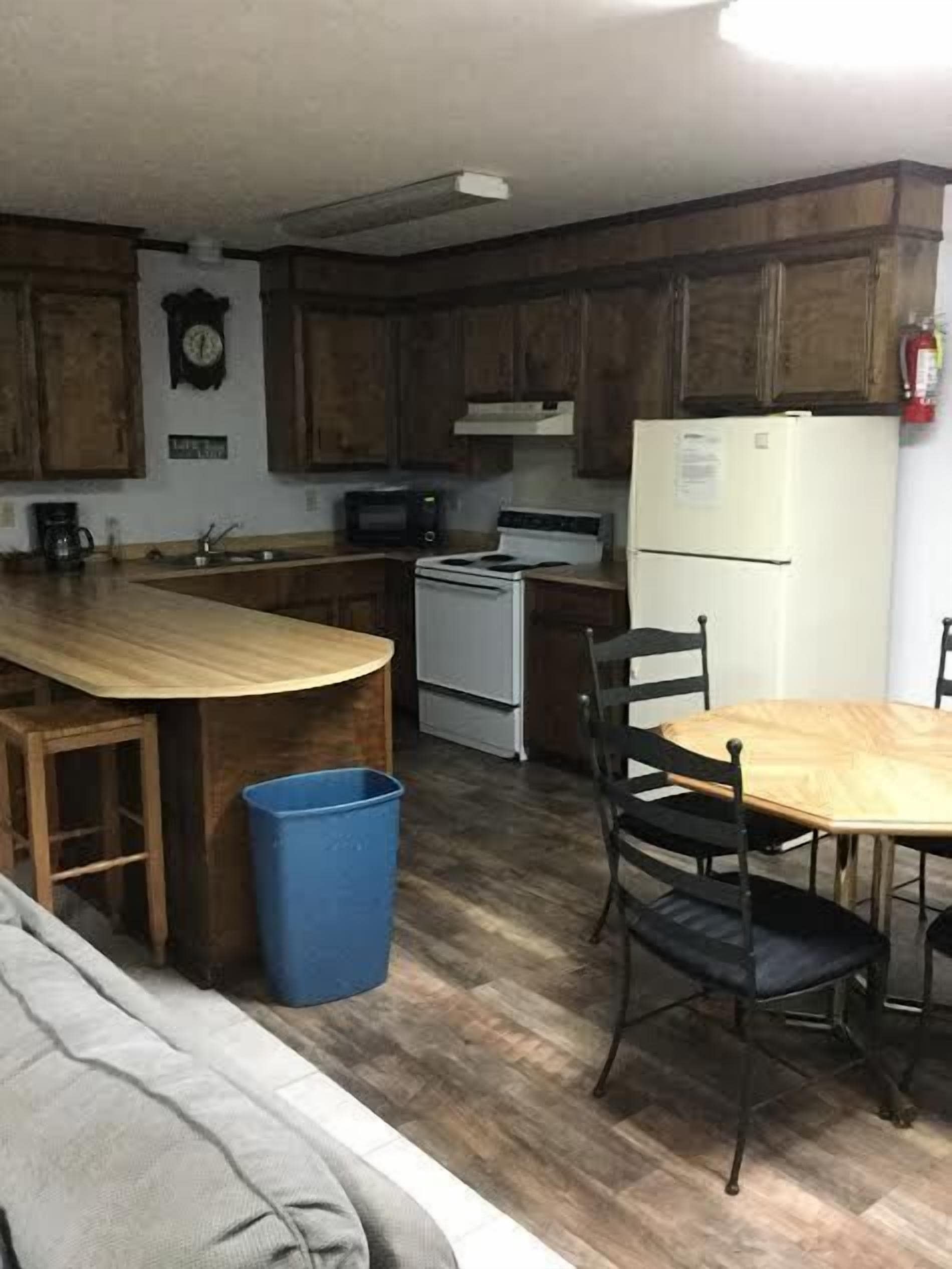 A compact kitchen with wooden cabinets, a microwave, a stove, and a refrigerator. There's a blue trash bin and a dining table with chairs.