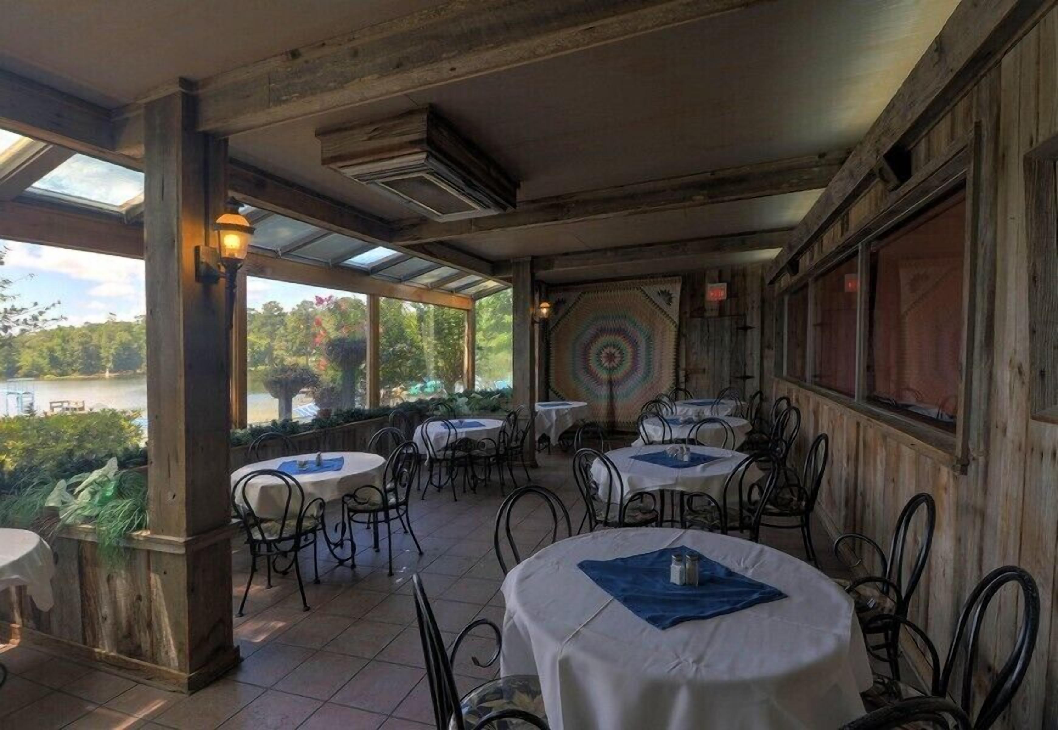 An outdoor dining area with round tables and chairs, a view of a lake, and a wooden wall with a decorative pattern.