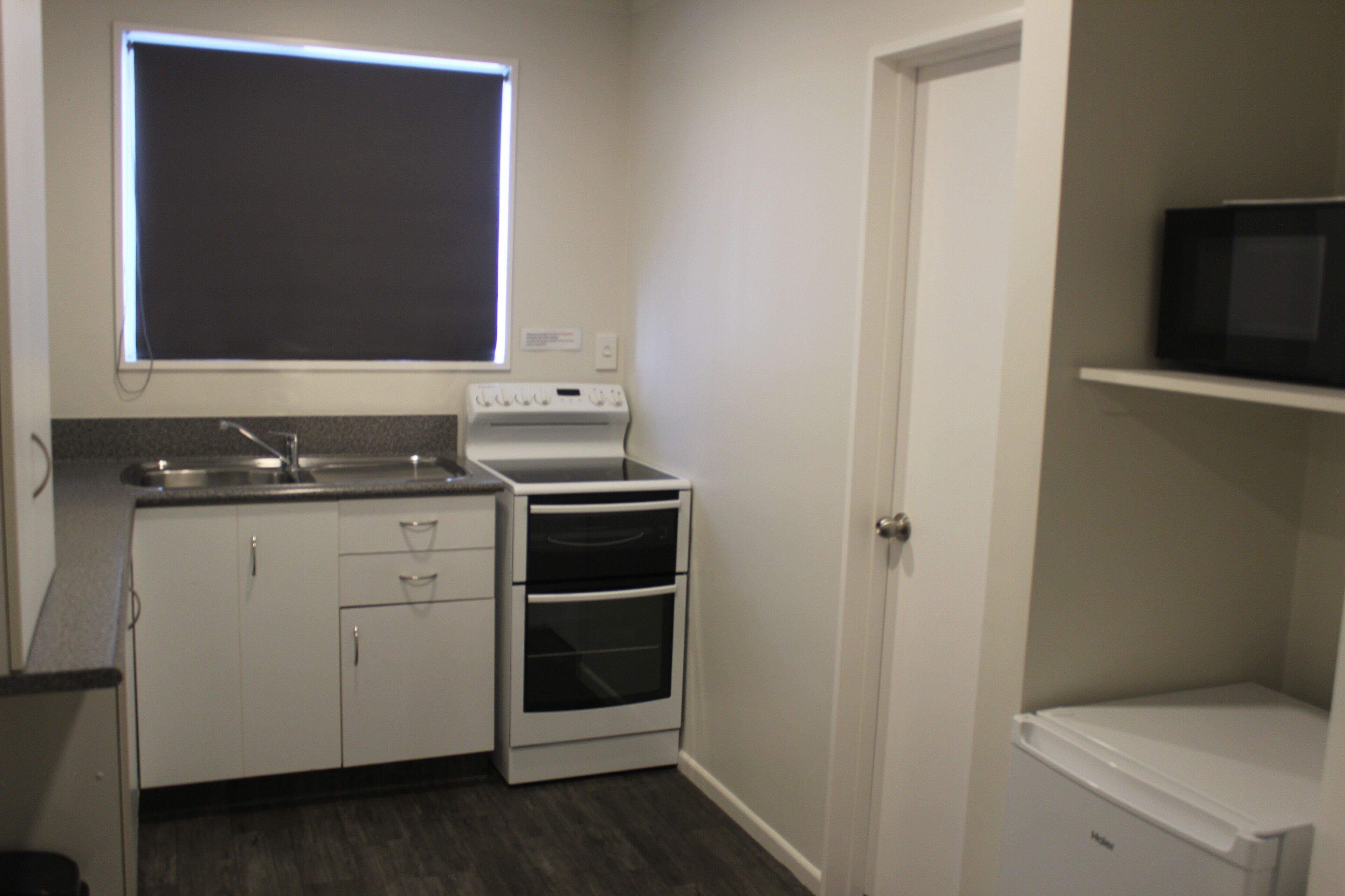 A compact kitchen with white cabinets, a built-in oven, a microwave on a shelf, and a window above the sink.