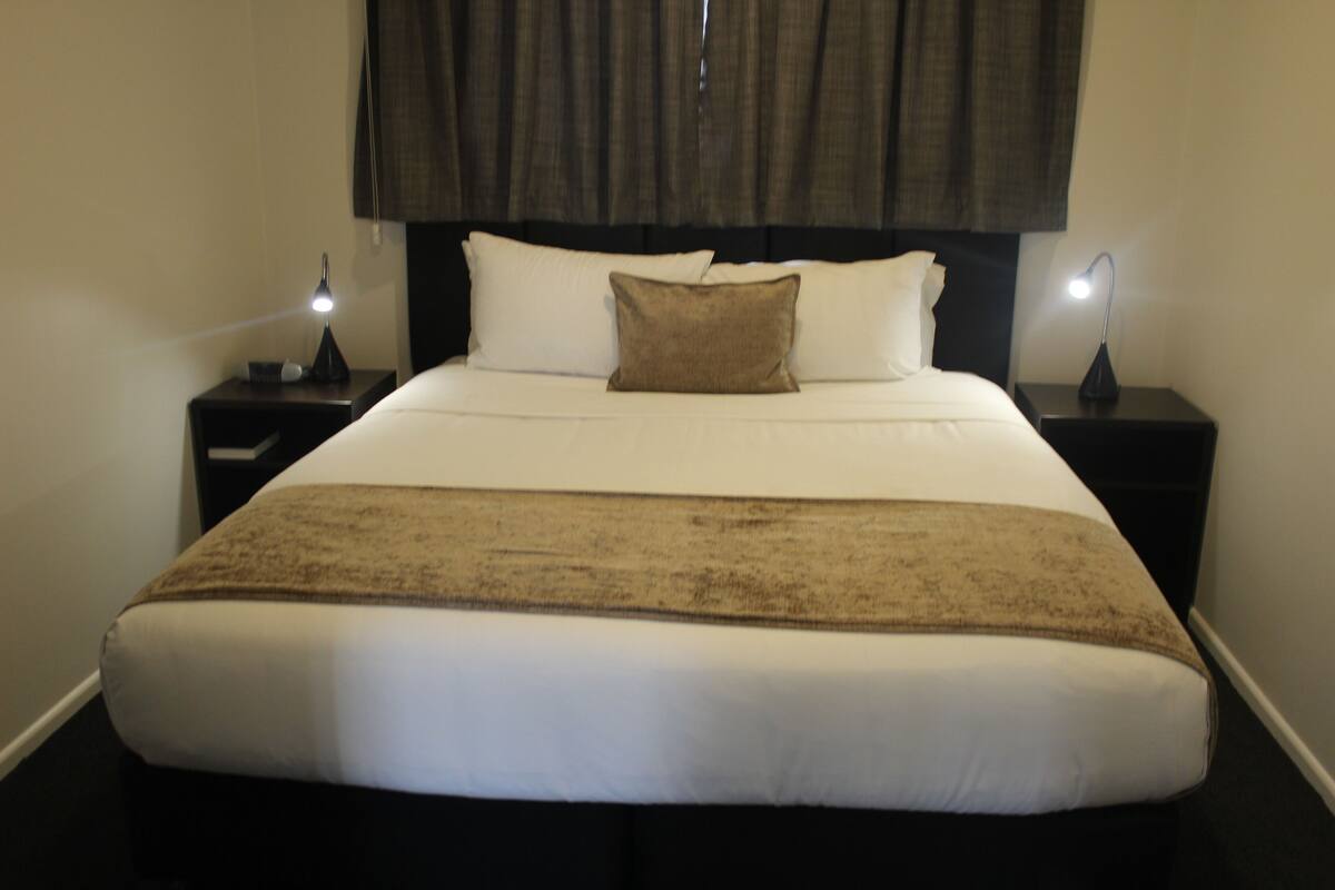 A neatly made bed with white linens and a brown throw blanket, flanked by two bedside tables with lamps.