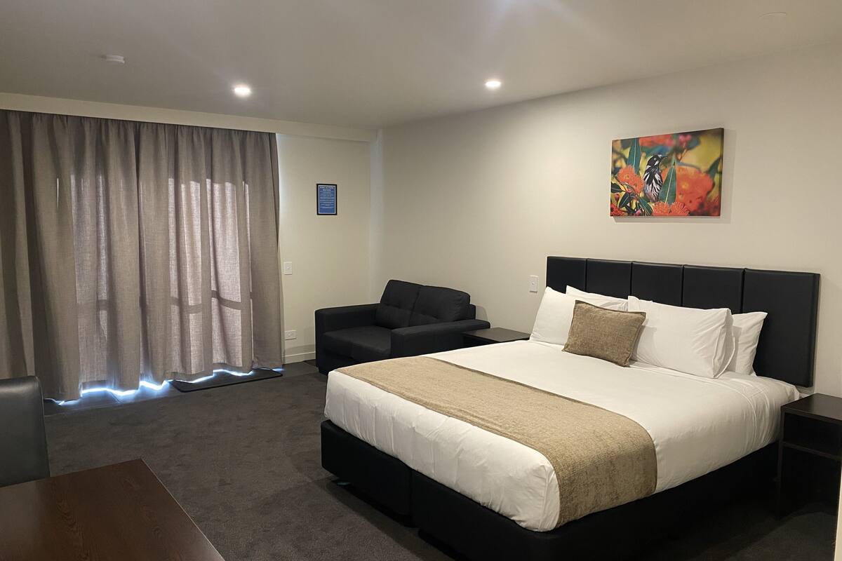 A hotel room with a large bed, a black headboard, a black sofa, a small table, and a painting on the wall.