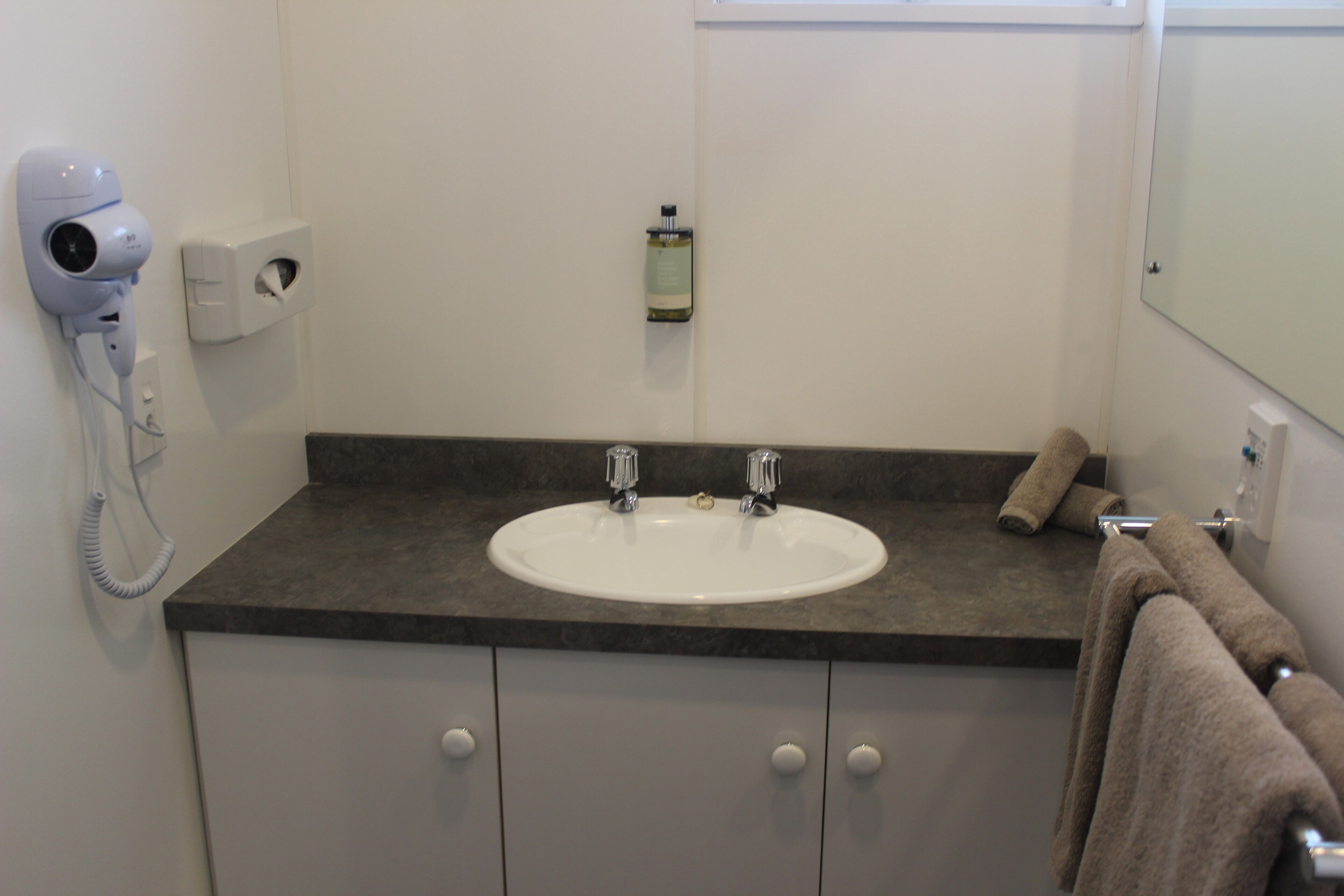 A bathroom with a white sink, two faucets, a towel rack with a towel, a hairdryer, and a soap dispenser on the wall.