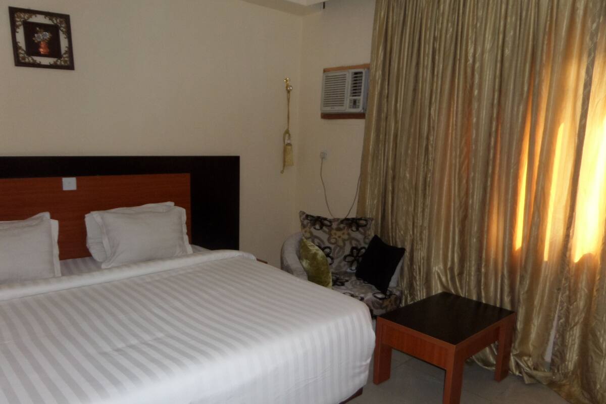 Una habitación de hotel con una cama, una mesita, una silla, un aire acondicionado de pared y una cortina.