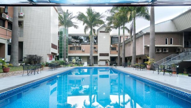 Une piscine dont le bord est revêtu de carrelage, entourée de palmiers et bordée d’un immeuble à plusieurs étages doté de balcons.