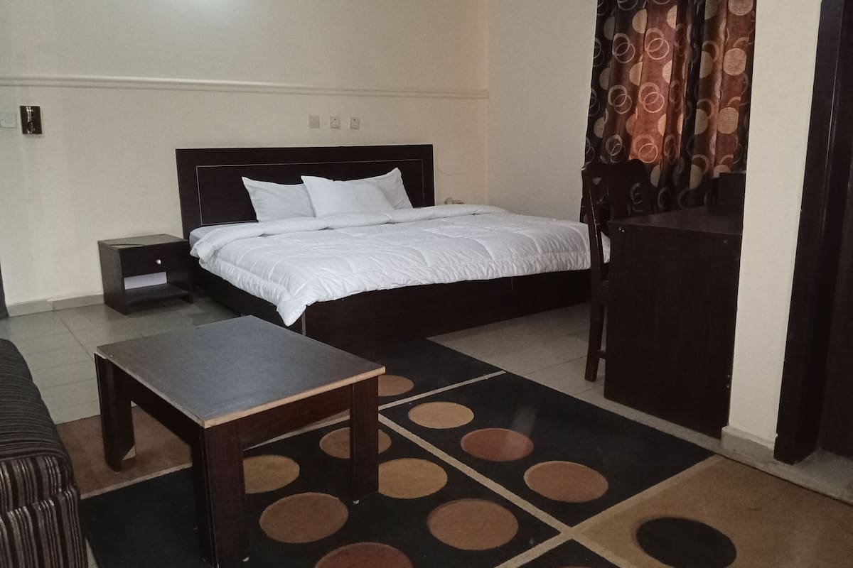 A hotel room with a bed, a nightstand, a table, and a patterned rug.
