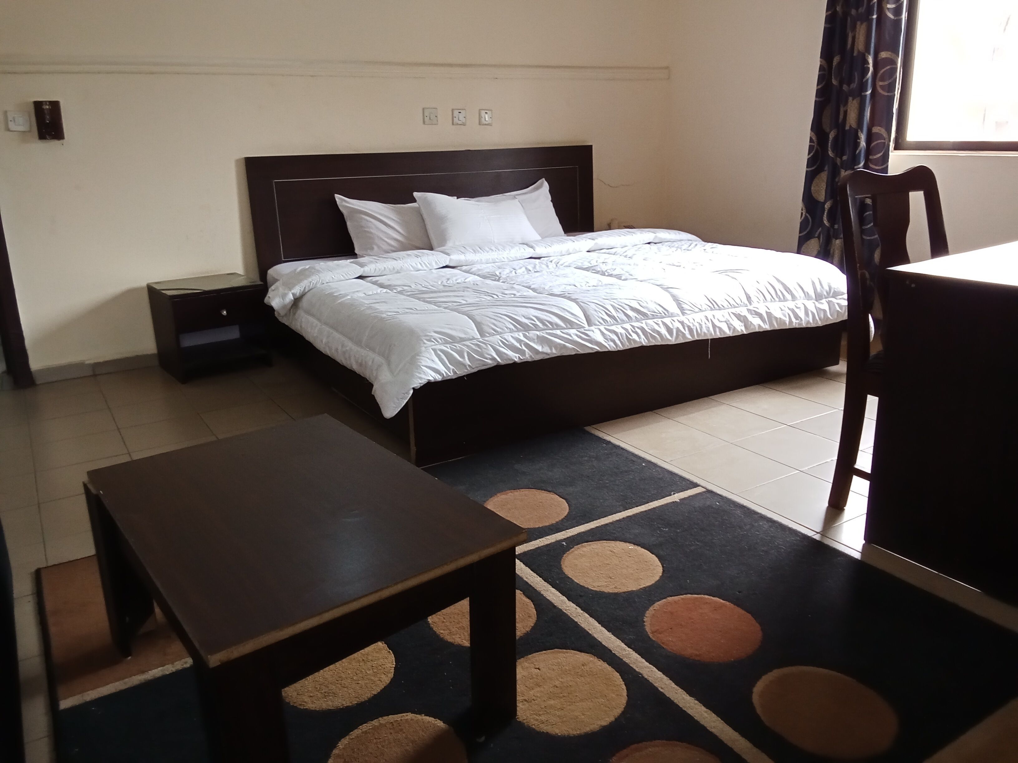 Una habitación de hotel con una cama, una mesita de noche, una mesa de madera y una alfombra estampada.