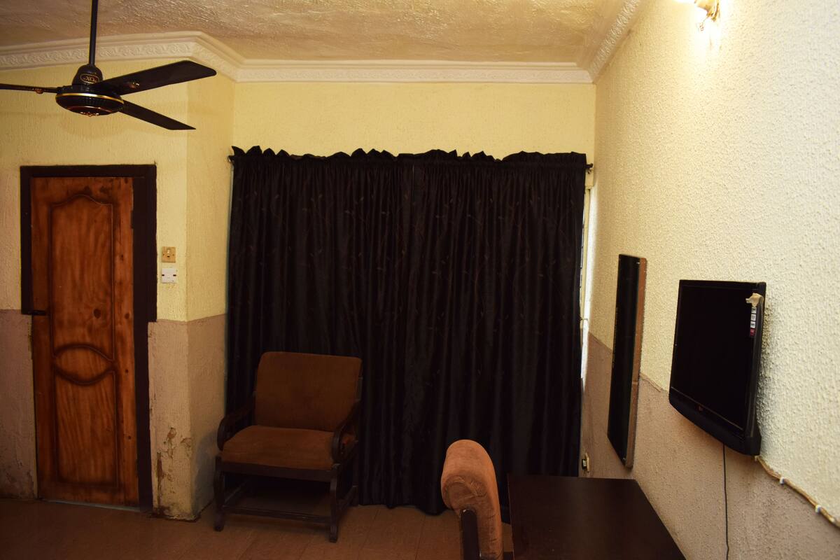 Quarto com ventilador de teto, porta de madeira, cortina escura, cadeira marrom e televisão na parede.