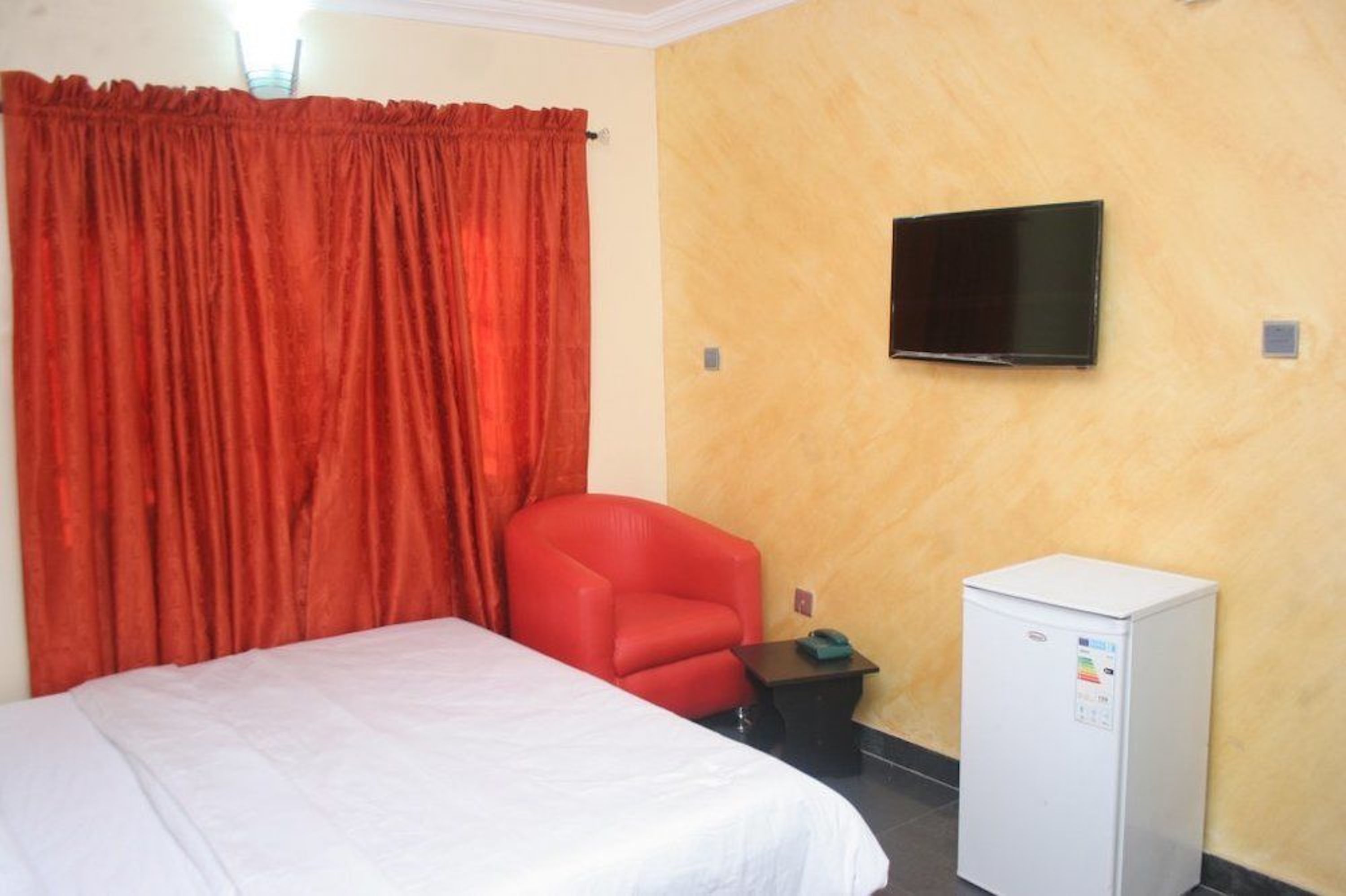 A hotel room with a bed, a red chair, a television on the wall, and a small white refrigerator.
