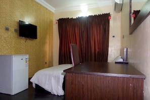 A room with a wooden desk, a television mounted on the wall, a small refrigerator, and a bed with a white sheet. - Blue Seasons Hotel & Suites (Lagos)