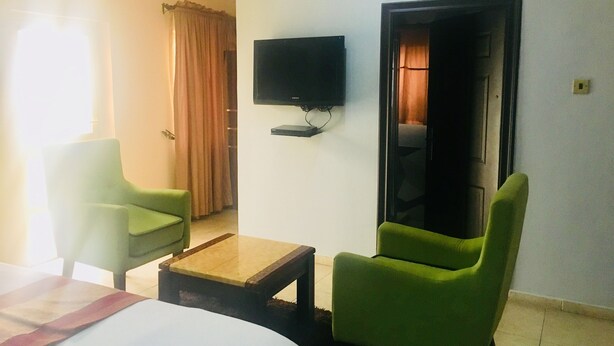 Habitación de hotel con una cama, dos sillones verdes, una mesita de madera, un televisor de pantalla plana en la pared y una ventana con cortinas.