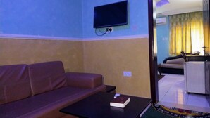 Una habitación con un sofá, una mesita con un libro, un televisor montado en la pared y vistas a otra habitación con una cama y un escritorio.