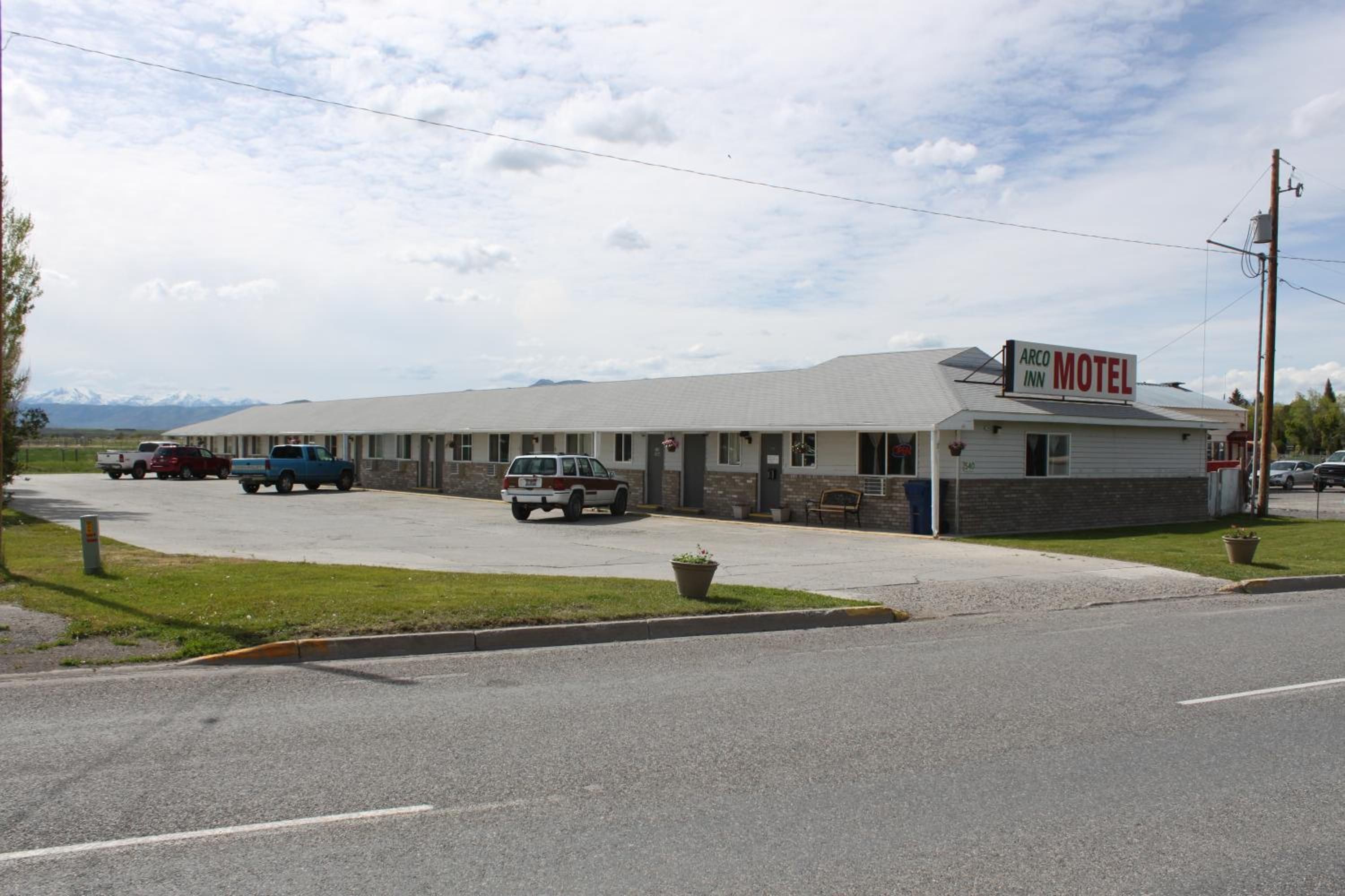 A motel with a simple, single-story structure, a parking lot with several vehicles, and a mountainous backdrop.