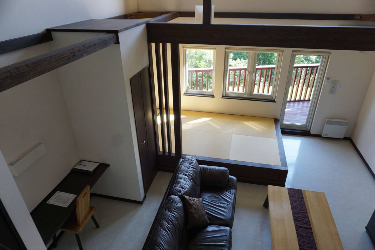 A modern living room with a dark leather sofa, a wooden coffee table, and a small desk with a chair. There are large windows with a view of a balcony.