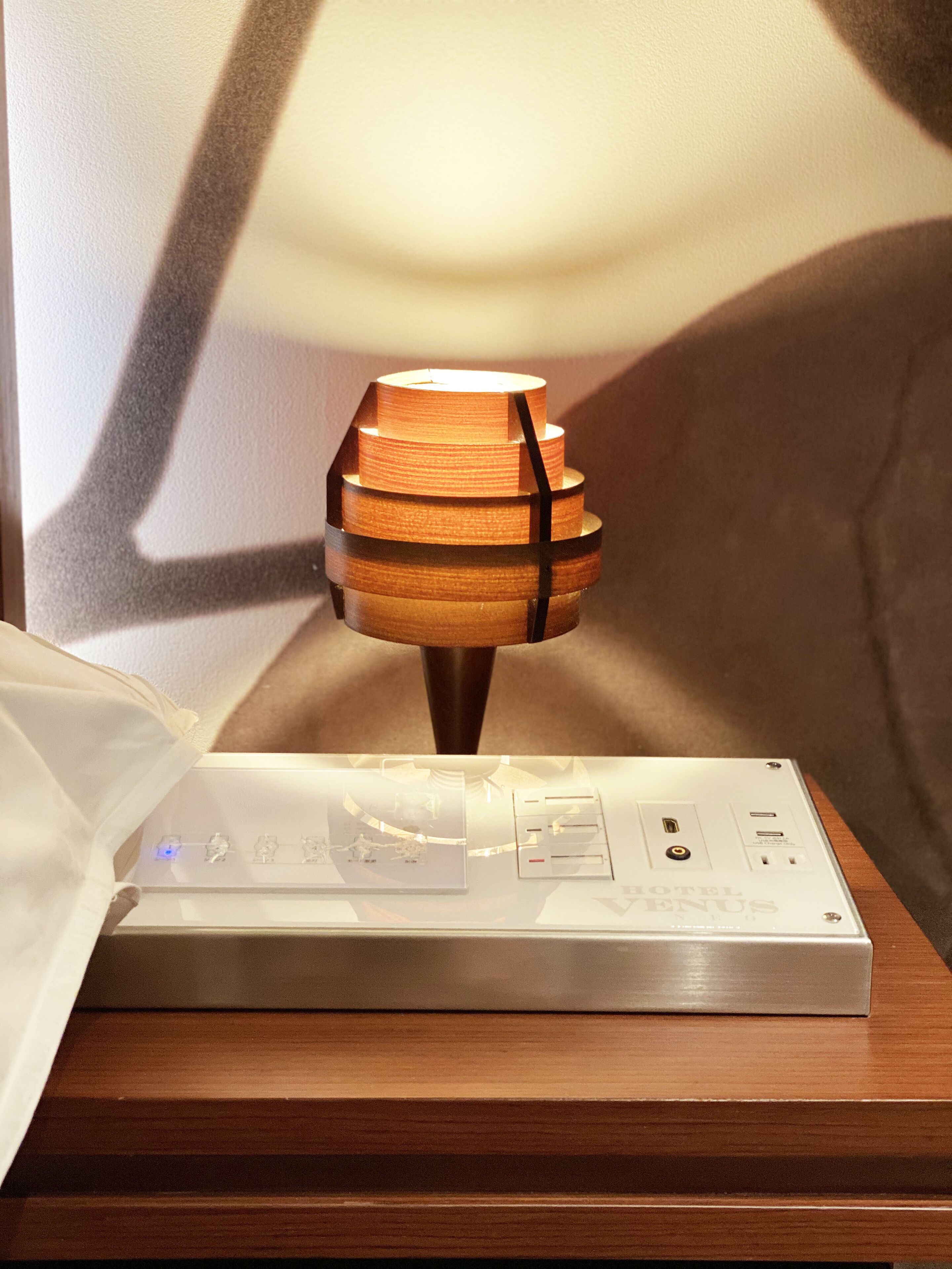 Una lámpara con base de madera y una banda negra, sobre una mesita de noche de madera junto a un dispositivo electrónico blanco con pantalla.