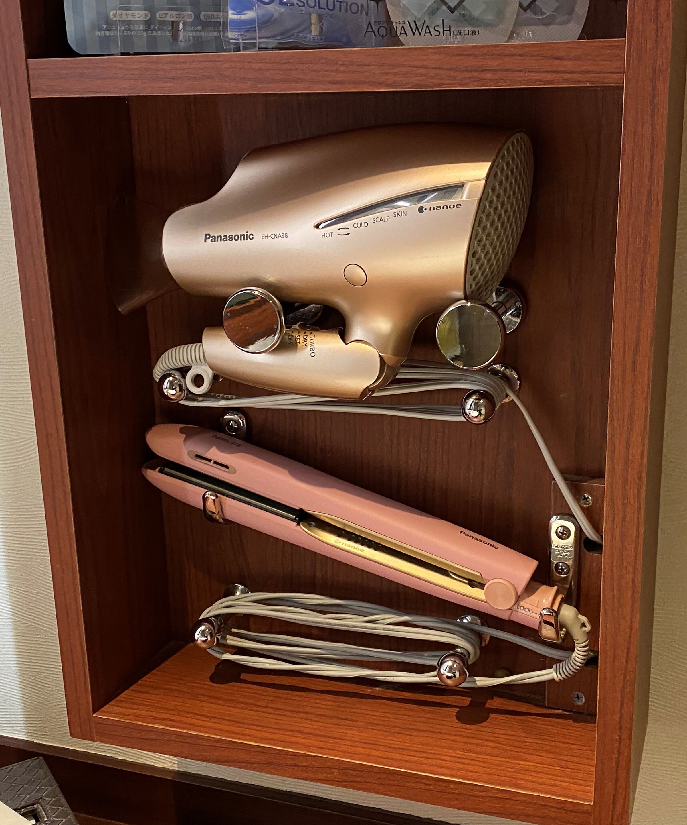 Un armario de madera con un secador de pelo Panasonic, un peine rosa y una plancha para el pelo dorada.